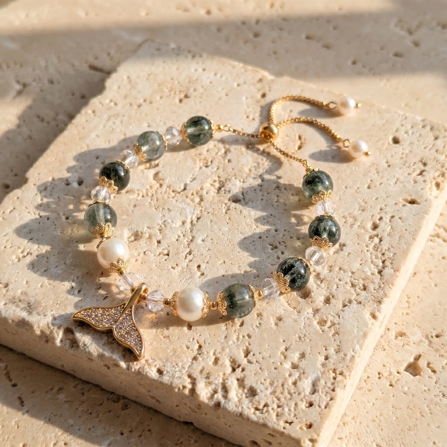 Flat lay of the Ocean Reverie bracelet coiled on a beige surface, emphasizing the sparkling fishtail charm and natural pearls.