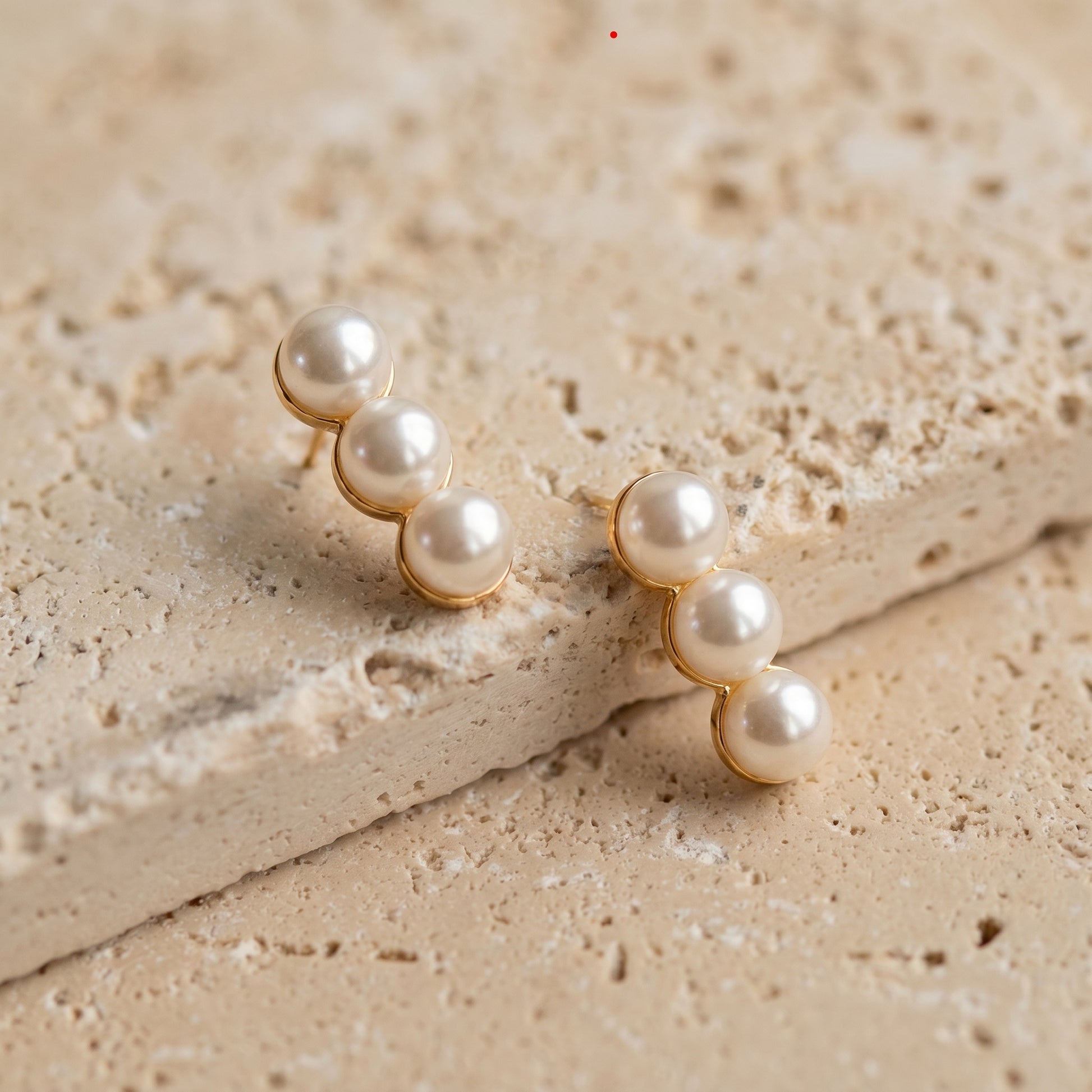 Dainty gold-plated linear earrings with three lustrous pearls laid flat on  a beige stone surface.