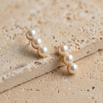 Dainty gold-plated linear earrings with three lustrous pearls laid flat on  a beige stone surface.