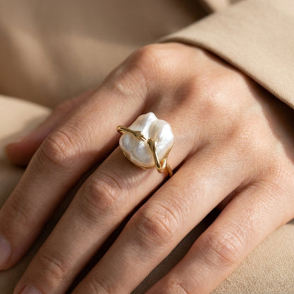 Close-up of The 'Embrace' ring on a finger, highlighting the gold hands holding a natural baroque freshwater pearl.