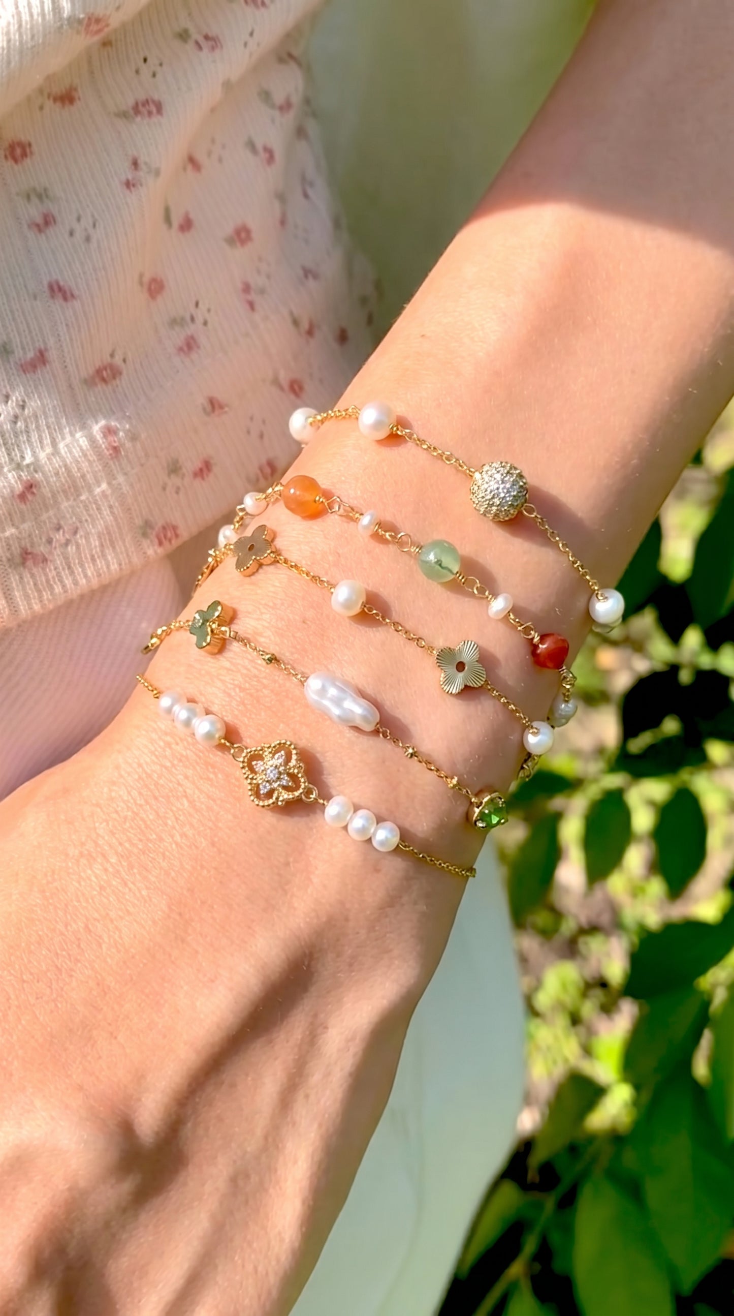 Close-up of a wrist wearing a stack of delicate 18K gold and natural pearl bracelets outdoors in the sunlight, featuring the Aurelia bracelet by Whisperland Studio against a green foliage backdrop.