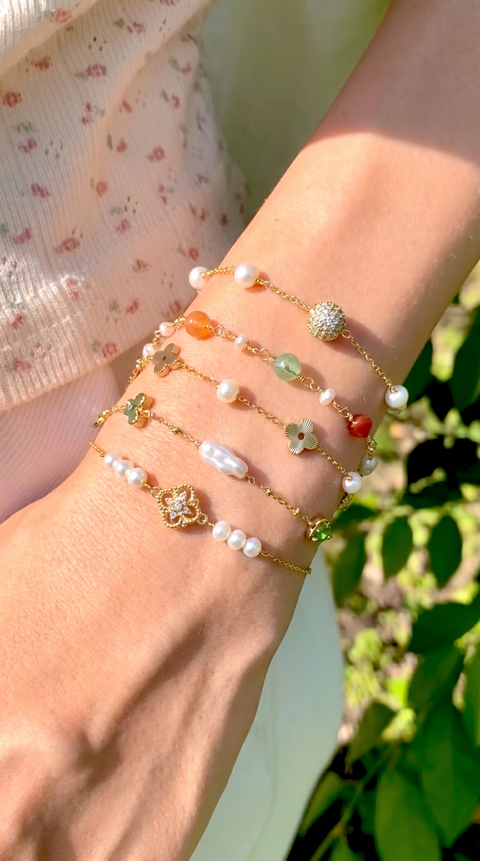 Close-up of a wrist wearing a stack of delicate 18K gold and natural pearl bracelets outdoors in the sunlight, featuring the Aurelia bracelet by Whisperland Studio against a green foliage backdrop.