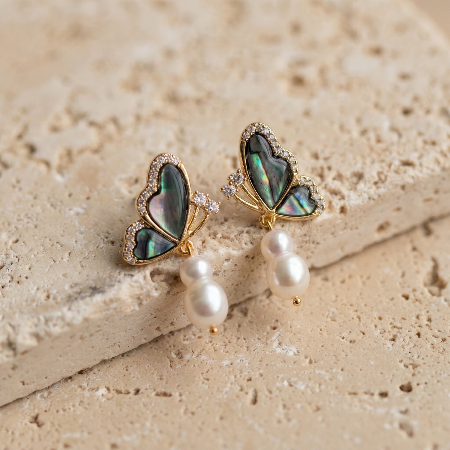 Nature-inspired minimalist earrings with abalone butterfly pendants resting on a textured stone background.