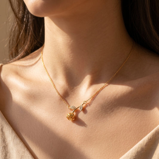 A close-up lifestyle shot of a delicate gold necklace with a small rose and leaf pendant, worn by a model in warm, direct sunlight.
