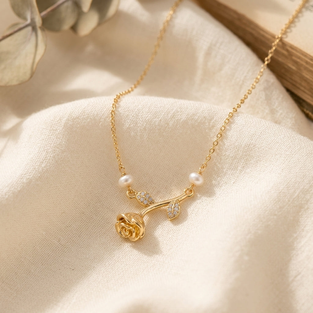 A delicate gold necklace with a rose-shaped pendant, a small pearl, and tiny clear stones, displayed on a soft cream fabric background.