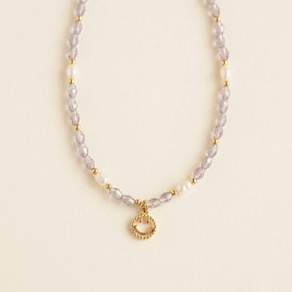 Whisperland Studio's 'Serotonin' necklace with purple crystal beads, unique freshwater pearls, and a happy face pendant, laid flat on a white background.