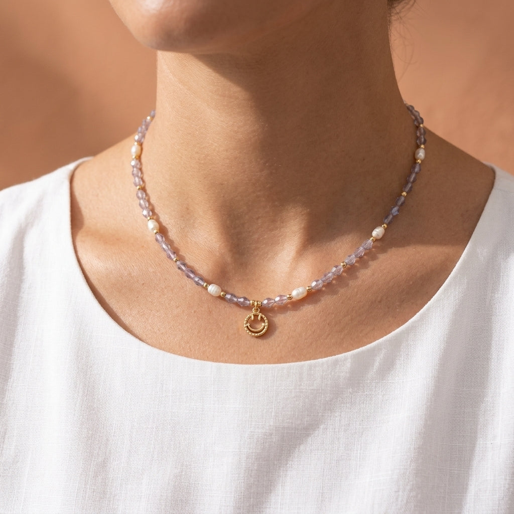The 'Serotonin' dainty smiley face necklace from Whisperland Studio, featuring pale purple crystal beads, natural freshwater pearls, and a 14K gold-plated happy face pendant on a model's neck.